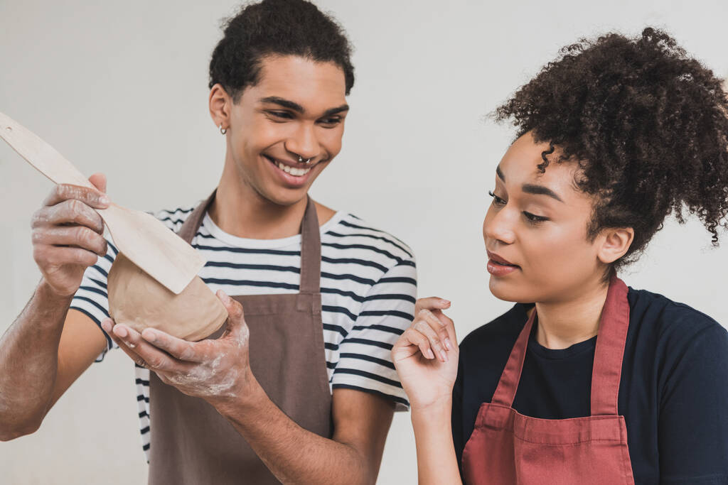 улыбающийся молодой африканский американец делает глиняный горшок с лопаткой рядом с женщиной с рукой рядом лицом в керамике - Фото, изображение