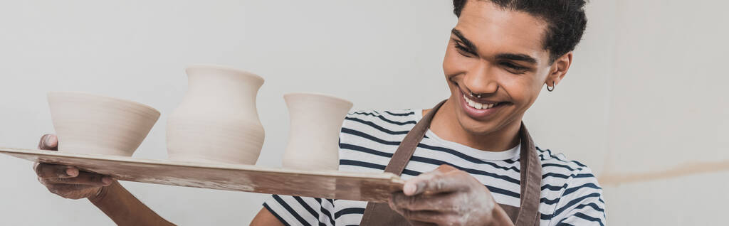 giovane uomo afroamericano sorridente guardando vasi di argilla su vassoio in ceramica, striscione - Foto, immagini