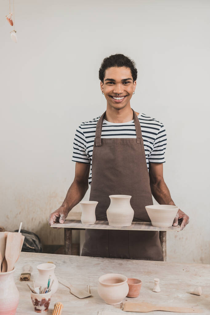 sorridente jovem afro-americano de pé com vasos de barro na bandeja em cerâmica - Foto, Imagem