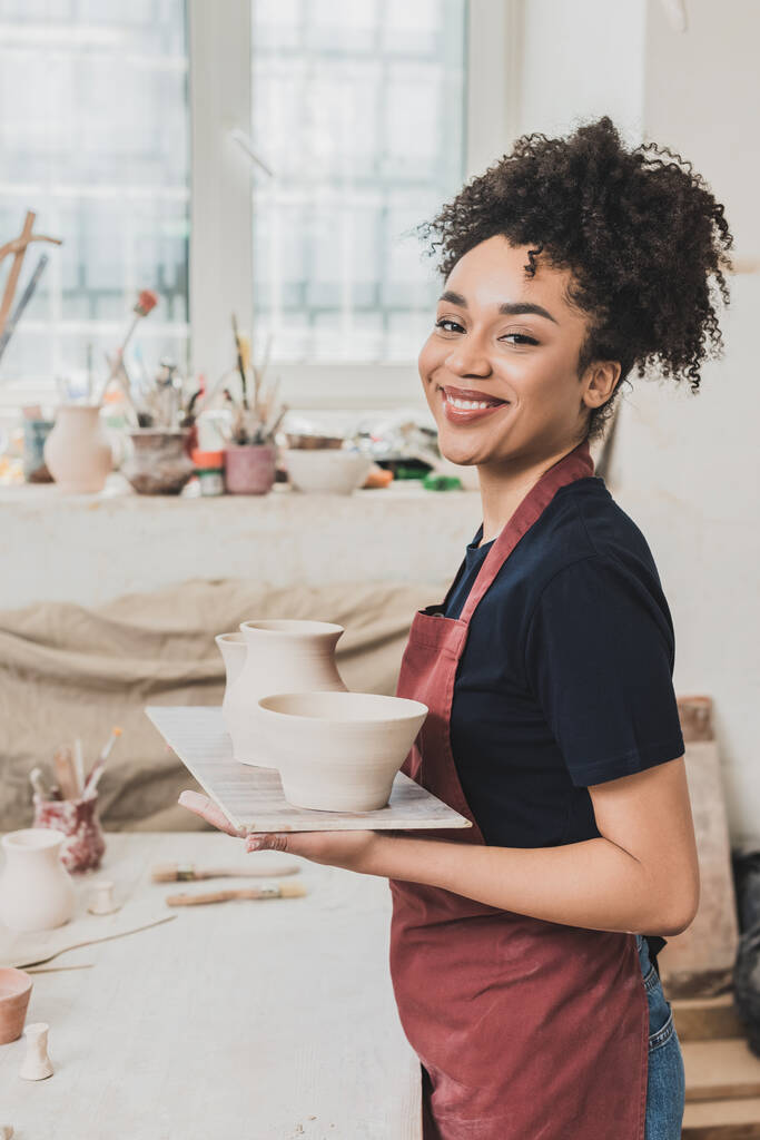 uśmiechnięta młoda Afroamerykanka trzymająca gliniane garnki na tacy w ceramice - Zdjęcie, obraz