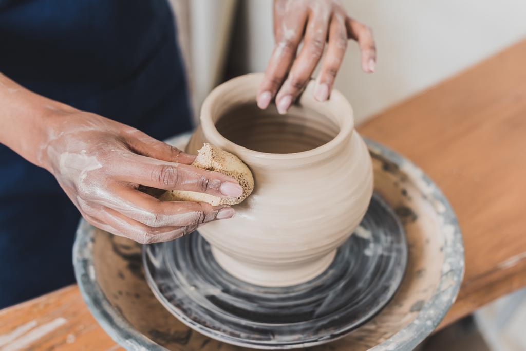 partial view of young african american woman modeling wet clay pot on wheel with hands and sponge in pottery - Fotoğraf, Görsel