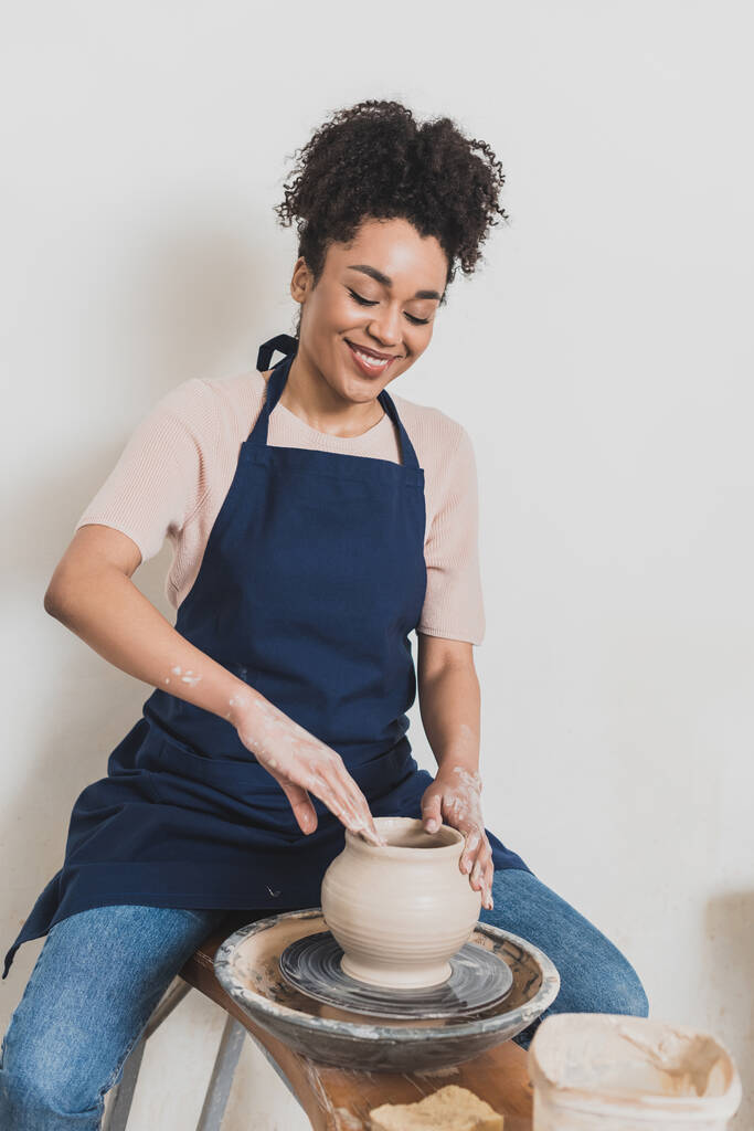 usmívající se mladá africká americká žena v ležérní oblečení a zástěra modelování mokrý hliněný hrnec na kole s rukama v keramice - Fotografie, Obrázek