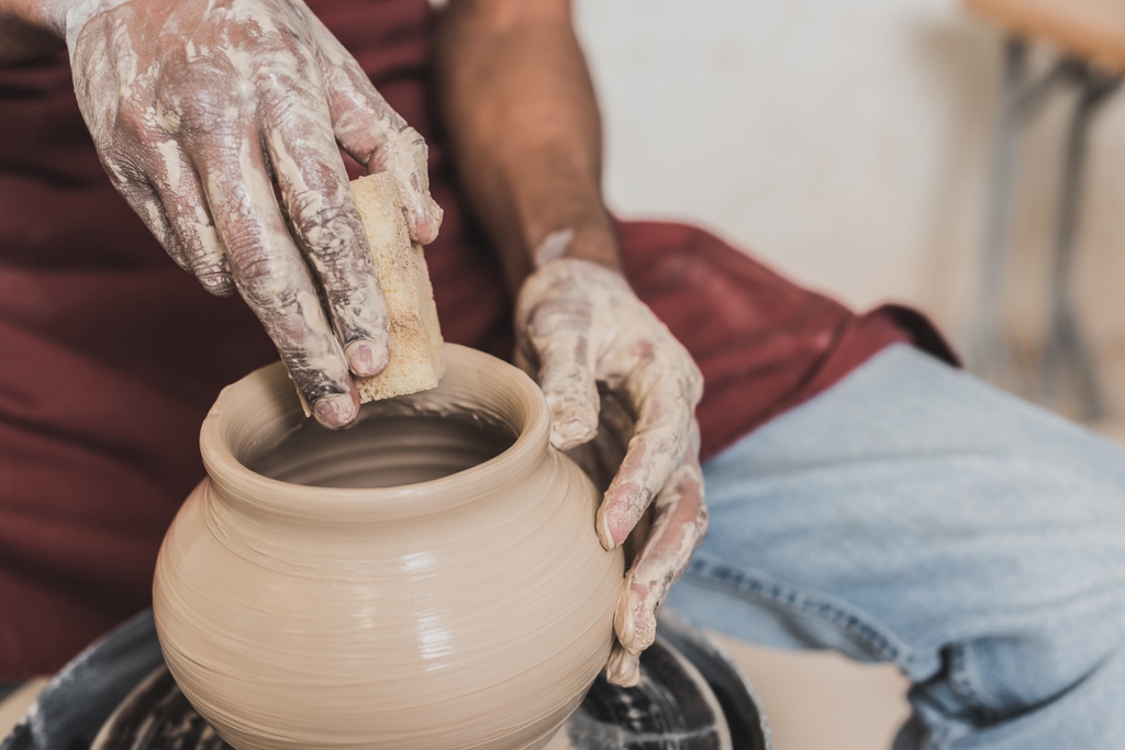 close up view of young african american man holding sponge and making wet clay pot on wheel in pottery - Fotoğraf, Görsel