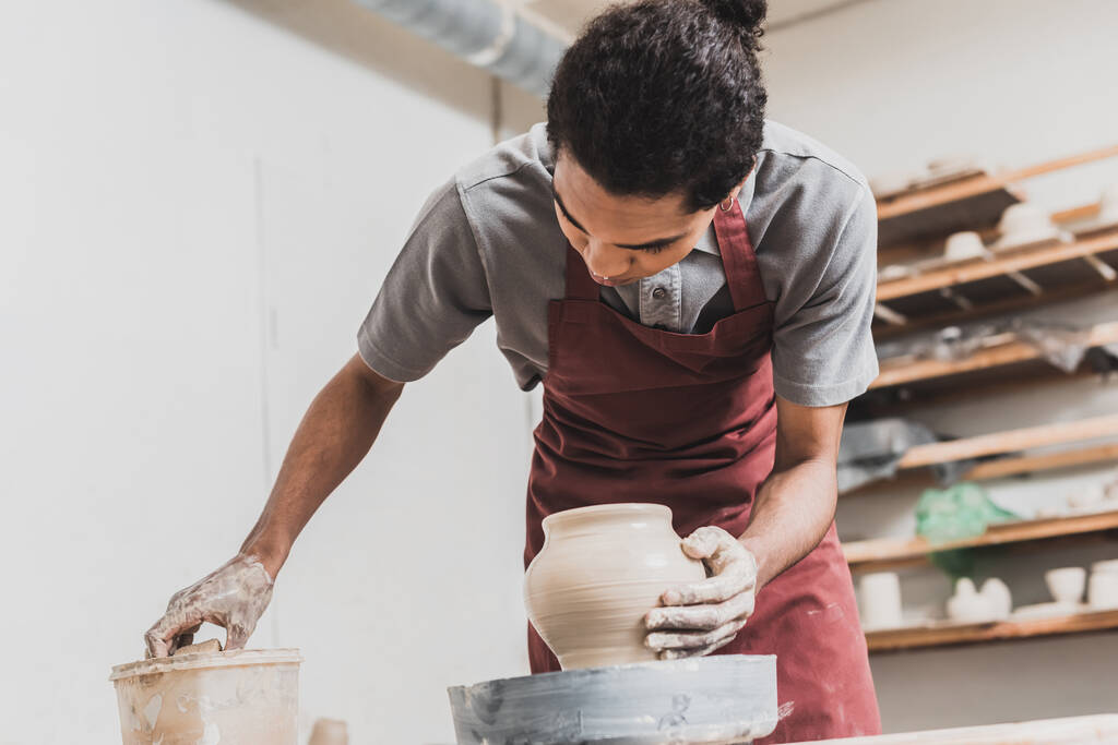 young african american man making wet clay pot on wheel and washing hand with sponge in plastic box in pottery - Photo, Image