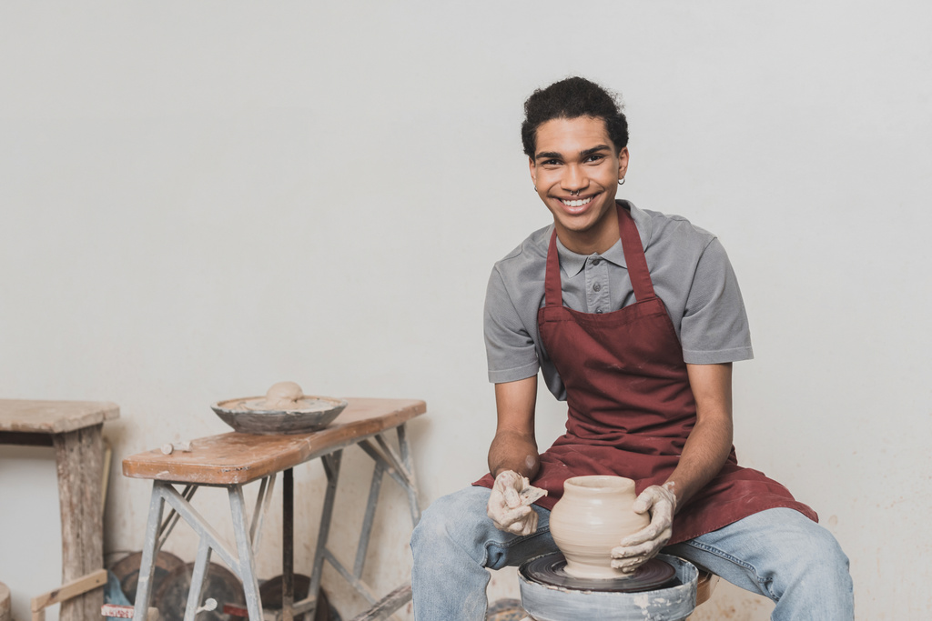 smiling young african american man shaping wet clay pot on wheel with scraper in pottery - Photo, Image