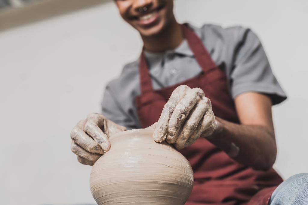 częściowy widok młodego Afroamerykanina człowieka w fartuchu rzeźbienie gliniany garnek w ceramice - Zdjęcie, obraz