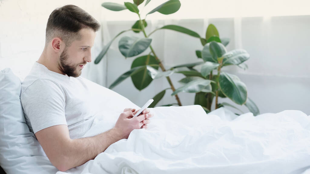 joven barbudo hombre usando teléfono inteligente en la cama - Foto, imagen