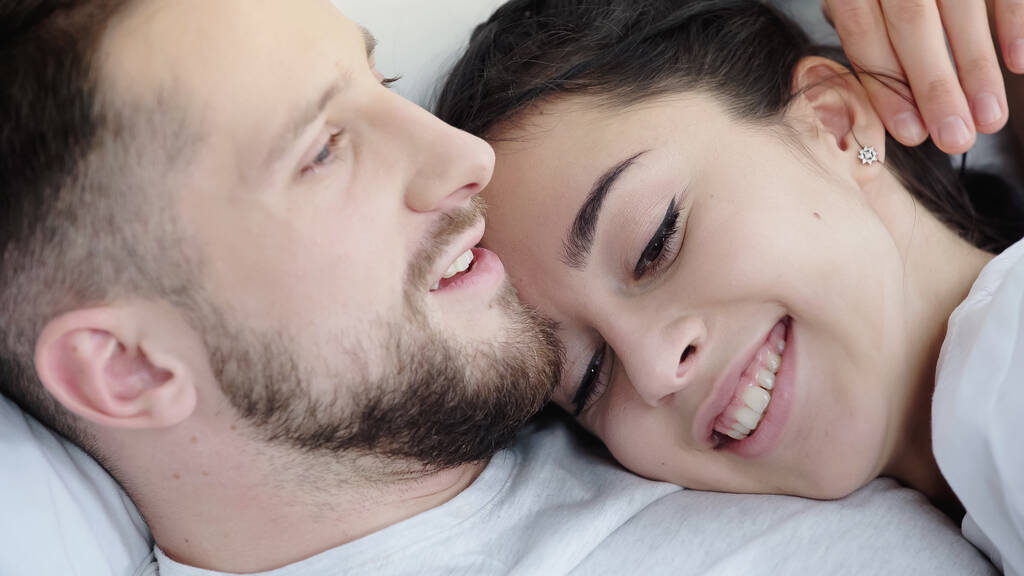 happy brunette woman leaning on bearded boyfriend while resting on bed - Photo, Image