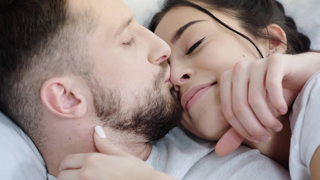 бородатый мужчина целует нос счастливой брюнетки, отдыхающей на кровати - Фото, изображение