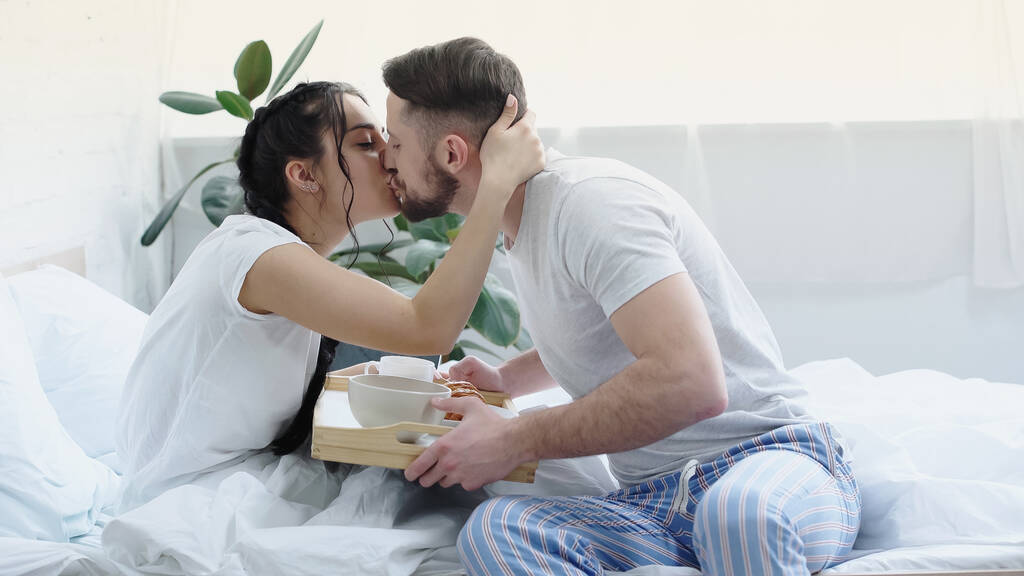 brodaty mężczyzna przynosząc śniadanie taca do dziewczyny z warkoczami podczas całowania w sypialni  - Zdjęcie, obraz