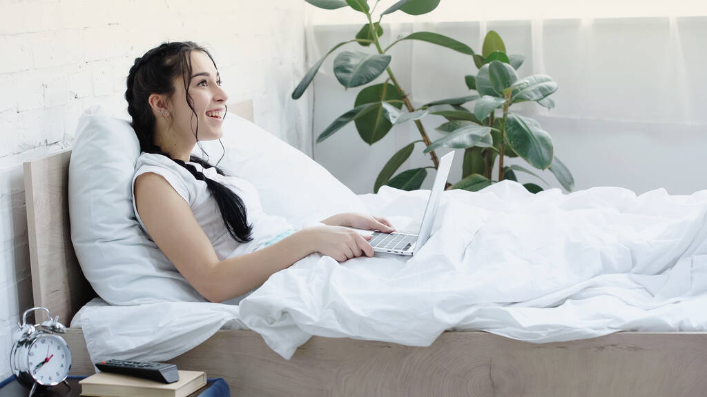 freelancer morena sorrindo enquanto usa laptop na cama - Foto, Imagem