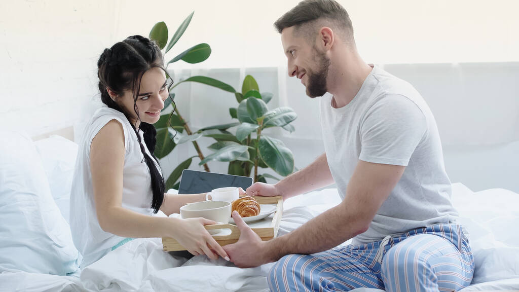 glücklicher Mann bringt Frühstückstablett zu freudiger Freundin mit Zöpfen im Schlafzimmer  - Foto, Bild
