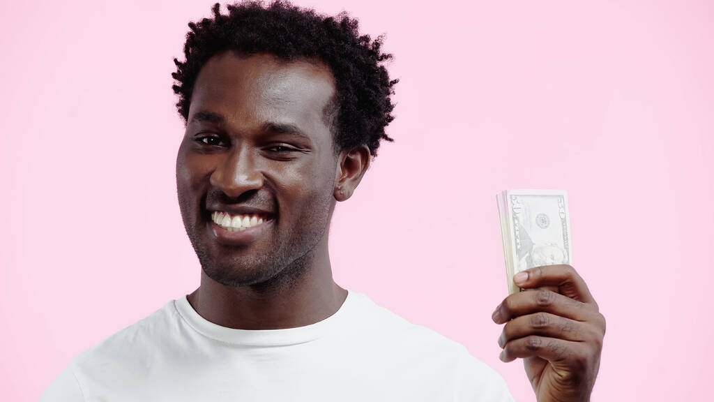 hombre afroamericano feliz en camiseta blanca sosteniendo dólares aislados en rosa - Foto, imagen