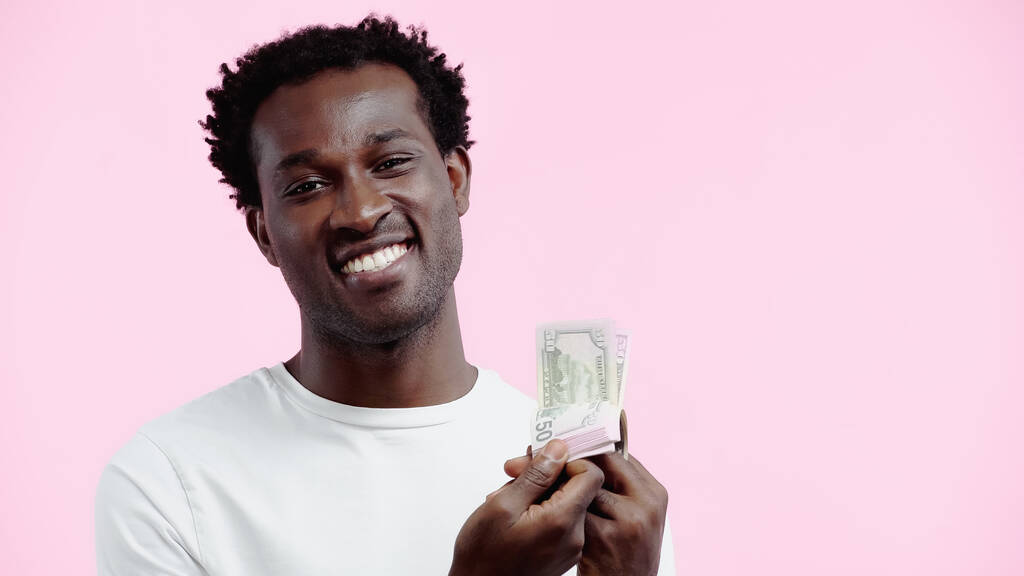 joyeux homme afro-américain en t-shirt blanc regardant la caméra tout en tenant des dollars isolés sur rose - Photo, image