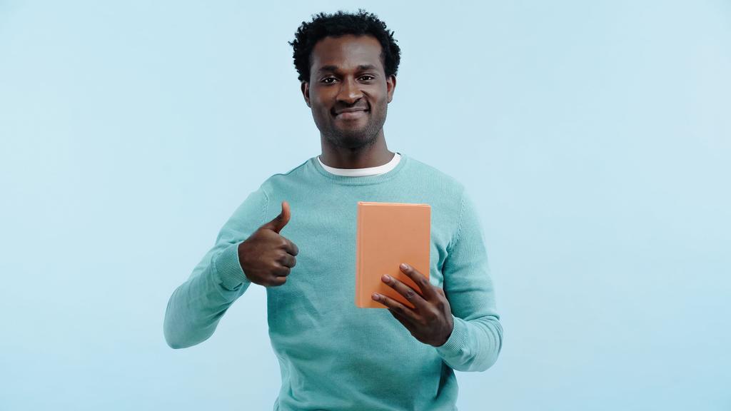 šťastný africký Američan drží knihu a ukazující palec nahoru izolované na modré - Fotografie, Obrázek