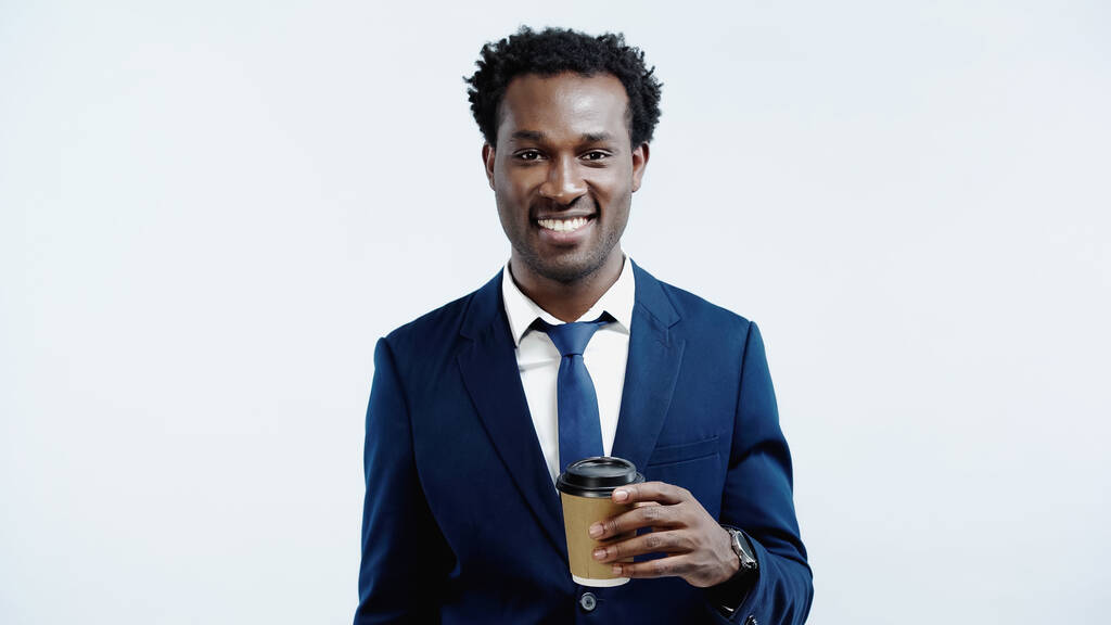 sonriente hombre de negocios afroamericano sosteniendo taza de papel con café para ir aislado en azul - Foto, imagen