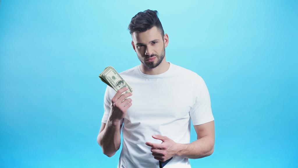 joven mirando a la cámara mientras muestra el dinero aislado en azul - Foto, imagen