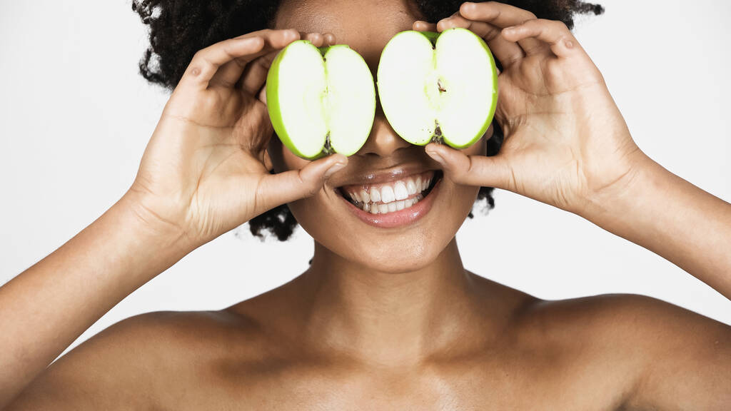 Smiling african american woman with naked shoulders holding pieces of apple near eyes isolated on grey  - Photo, Image