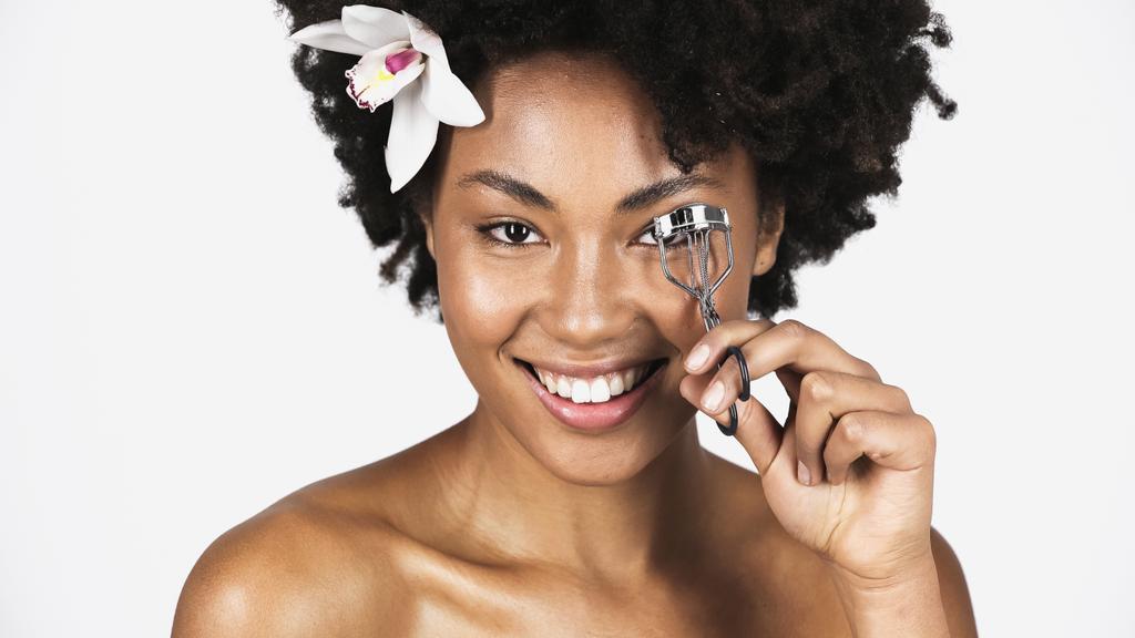 Uśmiechnięta Afroamerykanka z kwiatem we włosach przy użyciu lokówki do rzęs wyizolowanej na szaro  - Zdjęcie, obraz
