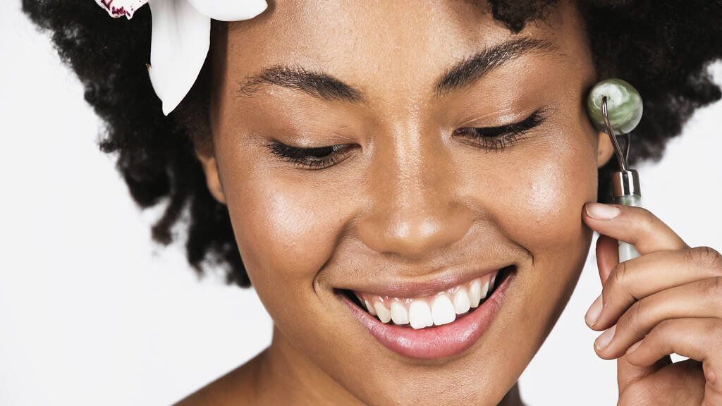 Close up άποψη του χαμογελαστού αφροαμερικανός γυναίκα με λουλούδι στα μαλλιά χρησιμοποιώντας νεφρίτη κυλίνδρου απομονώνονται σε γκρι  - Φωτογραφία, εικόνα