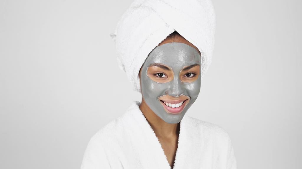 glimlachende Afro-Amerikaanse vrouw met handdoek op hoofd en gezicht masker geïsoleerd op grijs  - Foto, afbeelding