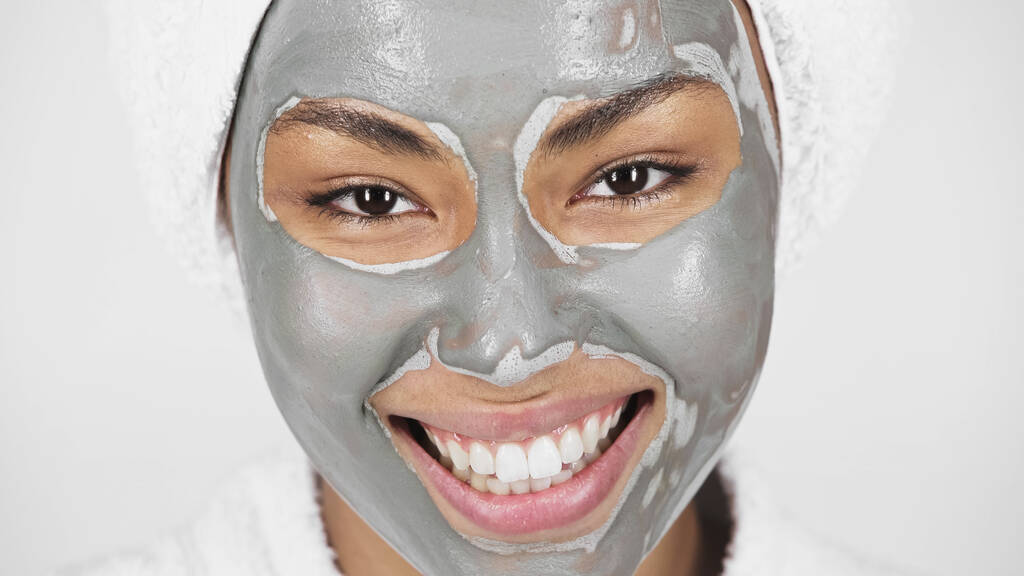 Στενή άποψη της χαρούμενης Αφροαμερικανής γυναίκας με πήλινη μάσκα στο πρόσωπο, απομονωμένη στο γκρι.  - Φωτογραφία, εικόνα