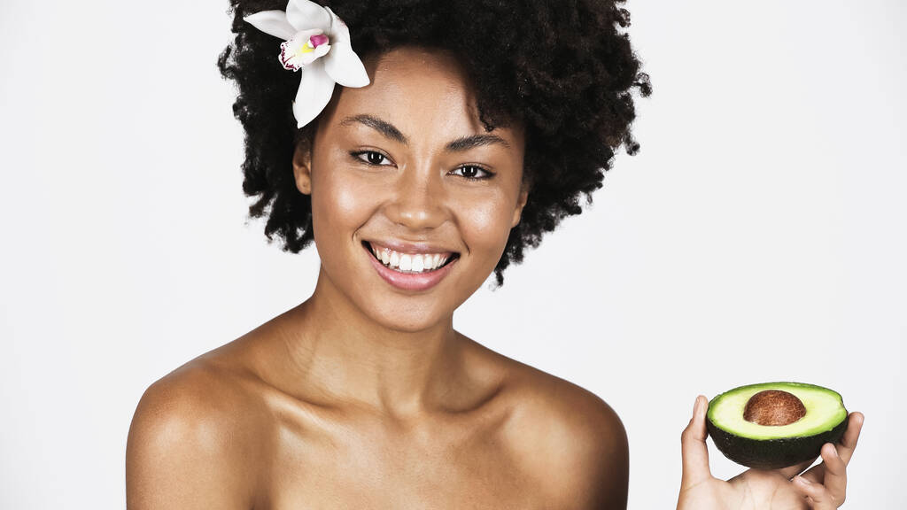 Wesoła Afroamerykanka z idealną skórą trzymająca połowę awokado na szarym  - Zdjęcie, obraz