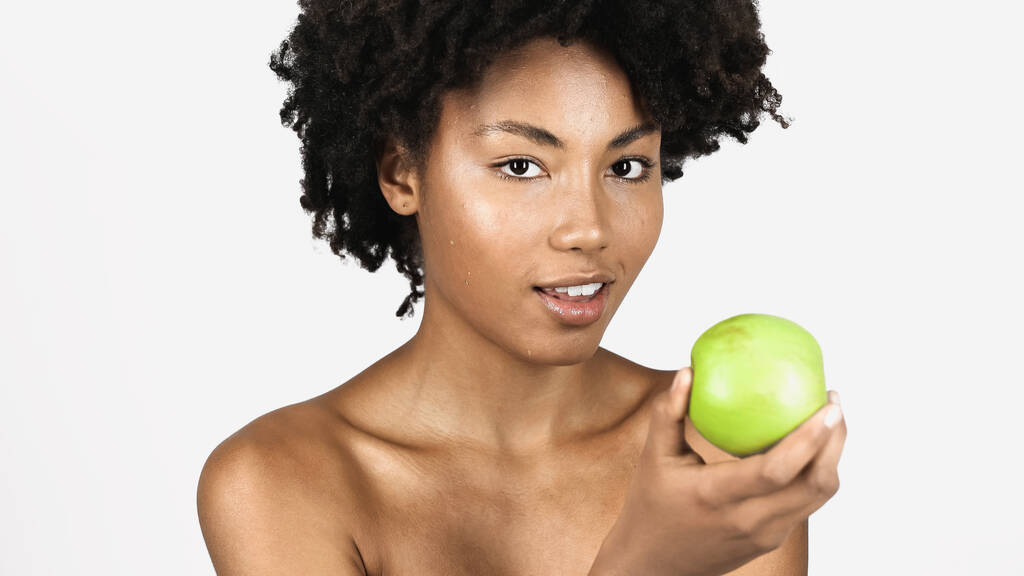 Довольно африканская американка с чистым лицом, держащая зеленое яблоко, изолированное на сером  - Фото, изображение