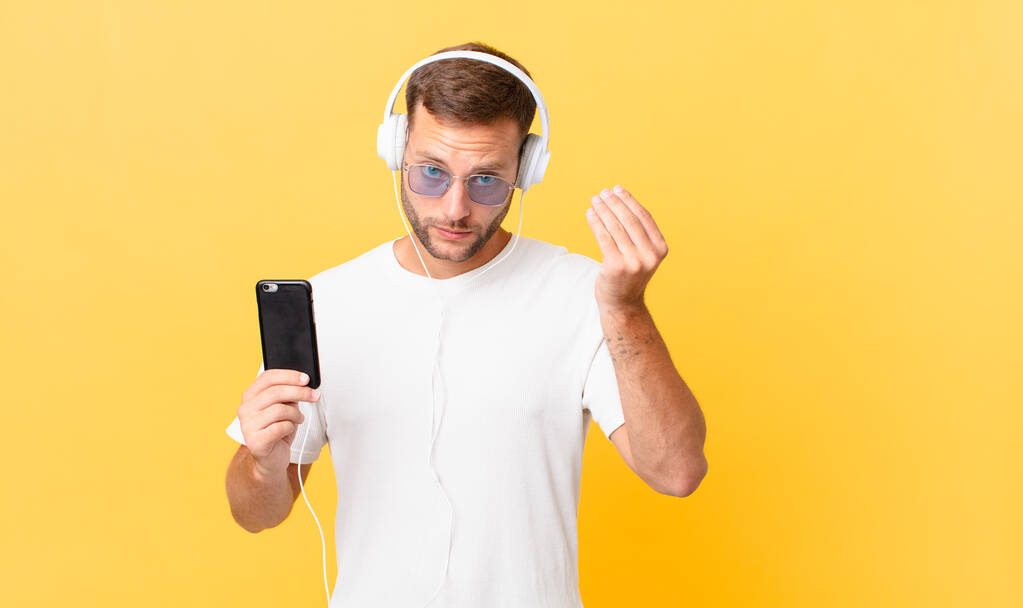 making capice or money gesture, telling you to pay, listening music with headphones and a smartphone - Фото, изображение