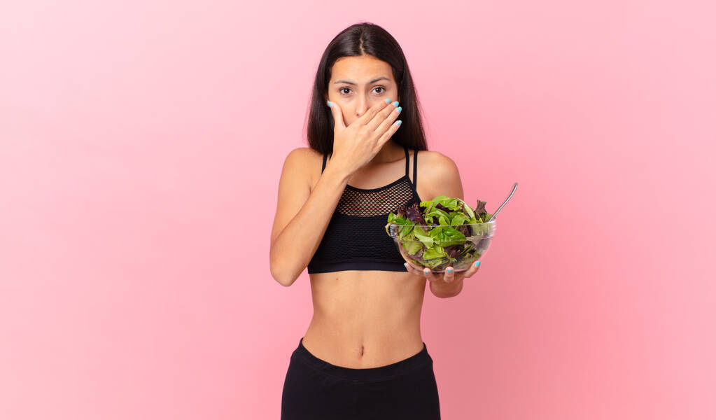 hispanic fitness woman covering mouth with hands with a shocked and holding a salad - Foto, afbeelding