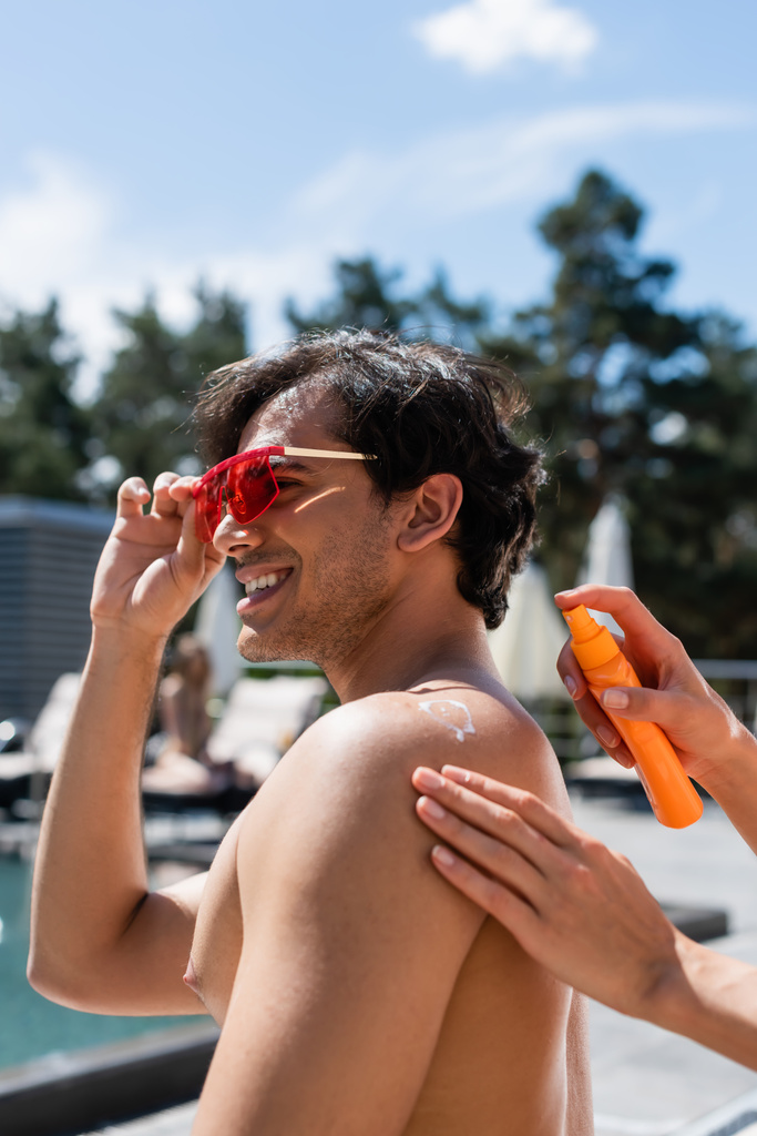 Frau trägt Sonnencreme auf Rückseite eines lächelnden Freundes mit Sonnenbrille auf - Foto, Bild