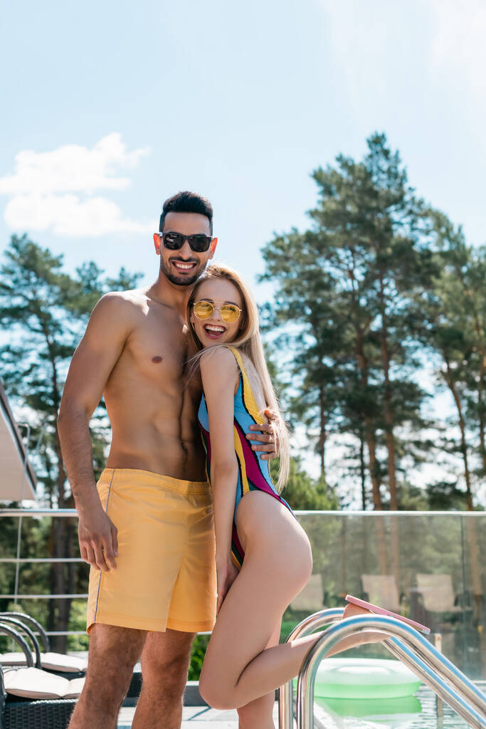 Arabský muž v plavkách objímá přítele v plavkách a slunečních brýlích u bazénu  - Fotografie, Obrázek