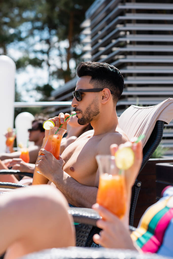 Arabo uomo in occhiali da sole bere cocktail vicino sfocato amico in costume da bagno  - Foto, immagini