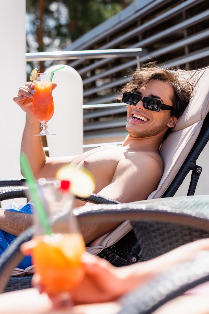 Uomo sorridente in occhiali da sole e costumi da bagno che tiene cocktail vicino amico sfocato durante il fine settimana  - Foto, immagini