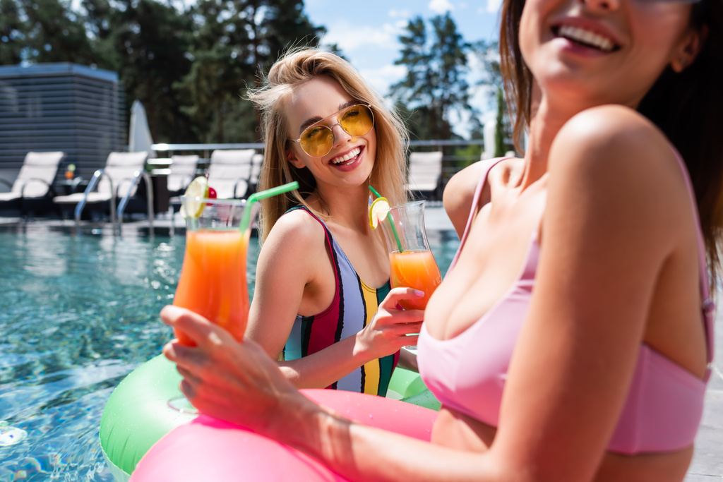 lachende vrouw in badpak en zonnebril houden cocktail in de buurt wazig vriend in het zwembad - Foto, afbeelding