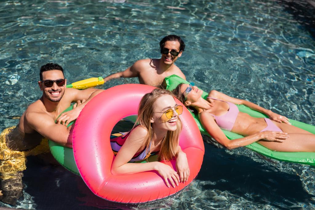 Mujer alegre riendo en el anillo de natación en la piscina cerca de amigos interracial - Foto, Imagen