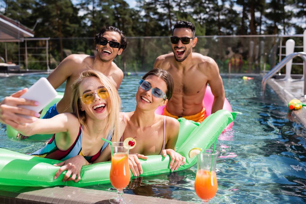 femme excitée prendre selfie dans la piscine près de cocktails et amis interracial - Photo, image