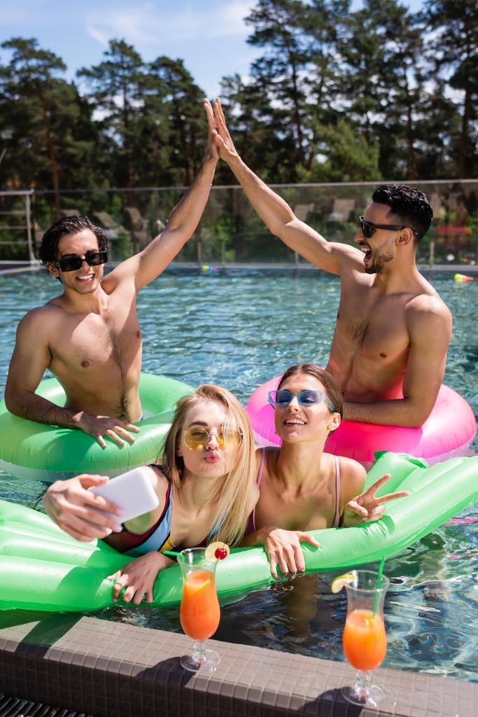 Mutlu ırklar arası erkekler yüzme havuzunda selfie çeken kadınlara beşlik çakıyor. - Fotoğraf, Görsel