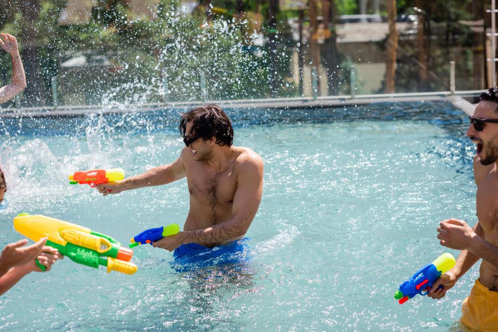 Aufgeregte multiethnische Freunde kämpfen mit Wasserpistolen im Schwimmbad - Foto, Bild