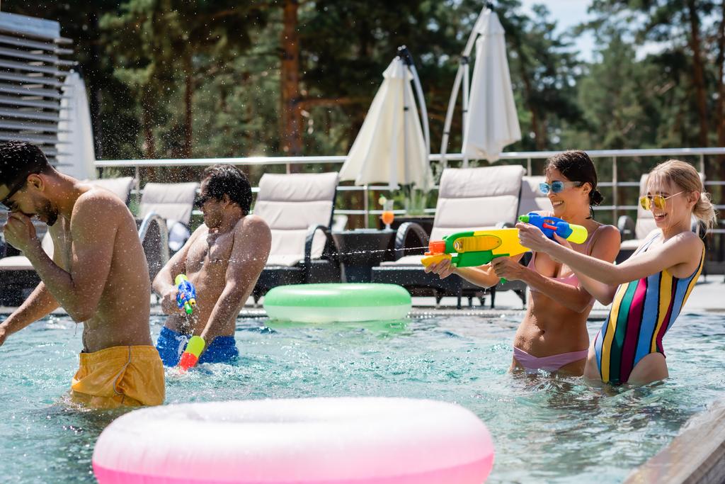 excited women fighting on water guns near multiethnic friends in swimming pool - Photo, Image