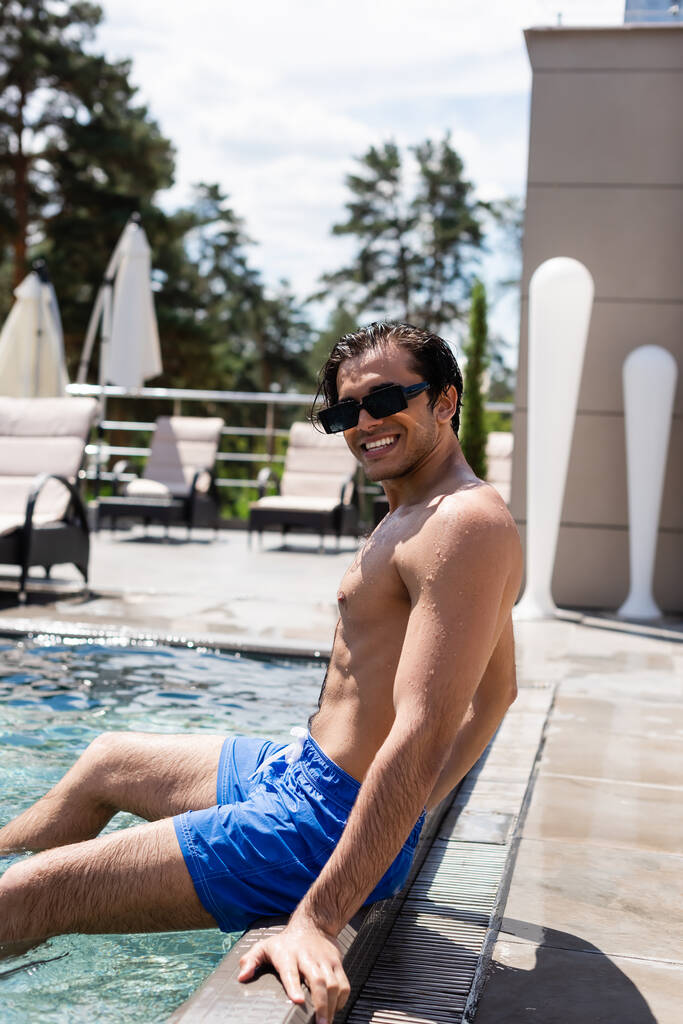 Hemdloser Mann in Badehose und Sonnenbrille lächelt am Pool - Foto, Bild