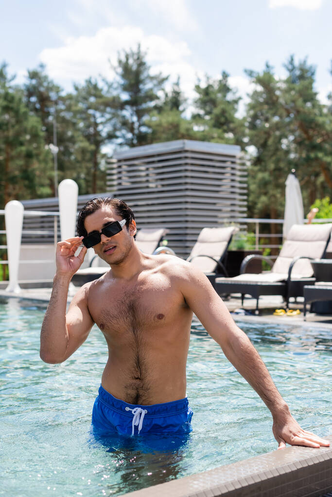 hombre sin camisa en traje de baño de pie en la piscina y el ajuste de las gafas de sol - Foto, imagen