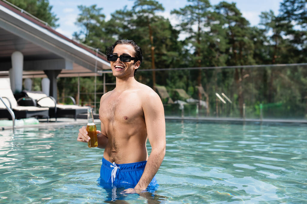 felice uomo senza maglietta in occhiali da sole rilassante in piscina con bottiglia di birra - Foto, immagini