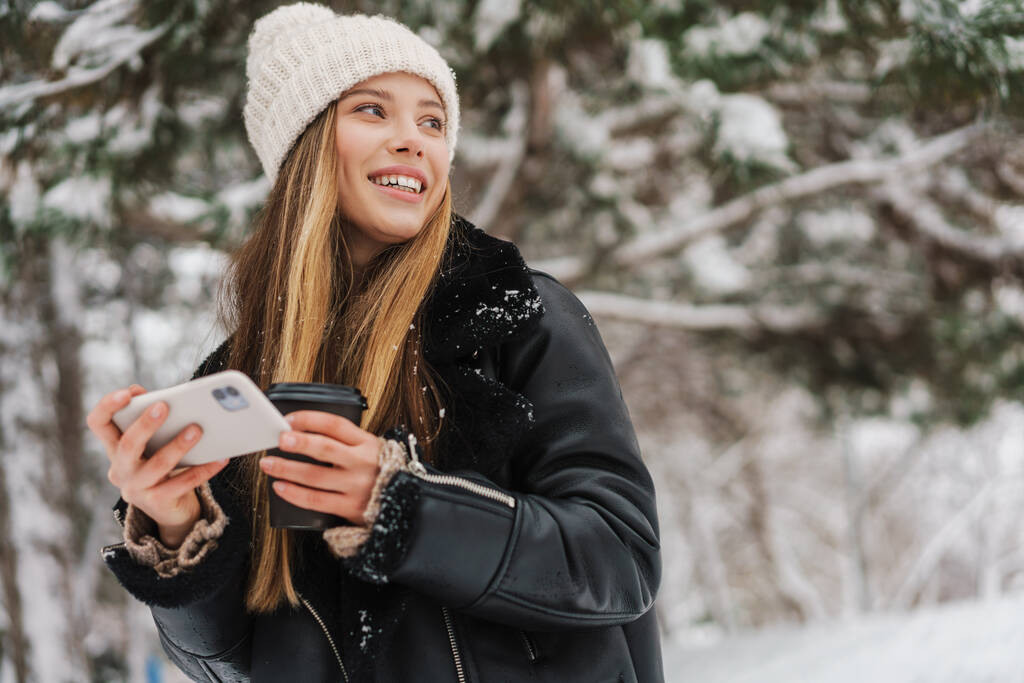 Mutlu kız, kış ormanlarında yürürken cep telefonu kullanıyor ve kahve içiyor. - Fotoğraf, Görsel
