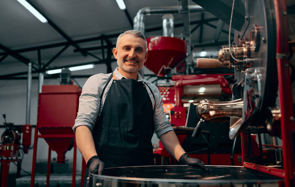 Πορτρέτο του χαμογελαστού καφέ roaster στέκεται στο ψήσιμο μηχάνημα και κοιτάζοντας την κάμερα. Επιλεκτική εστίαση. - Φωτογραφία, εικόνα