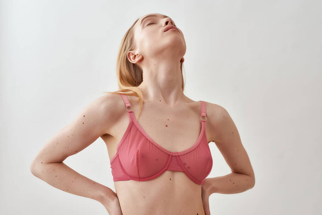 Sensual slim young caucasian woman wearing pink transparent bra lifting her head up while posing with eyes closed isolated over light gray background - Foto, Imagem