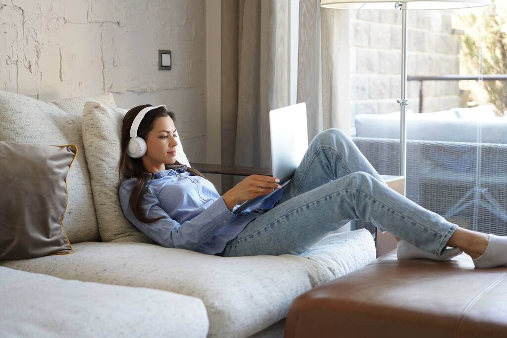 Chica bonita usando el ordenador portátil en el sofá en casa en la sala de estar. Escuchar música, relajarse después de una semana difícil - Foto, imagen