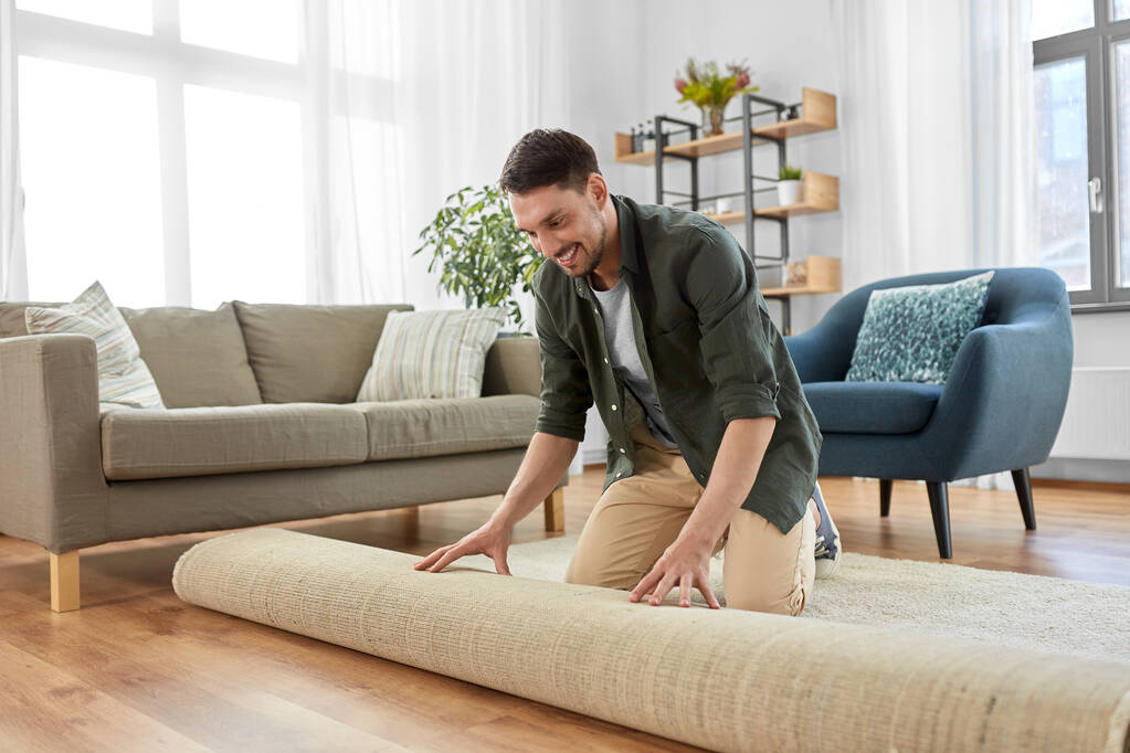 щасливий усміхнений молодий чоловік розгортає килим вдома
 - Фото, зображення