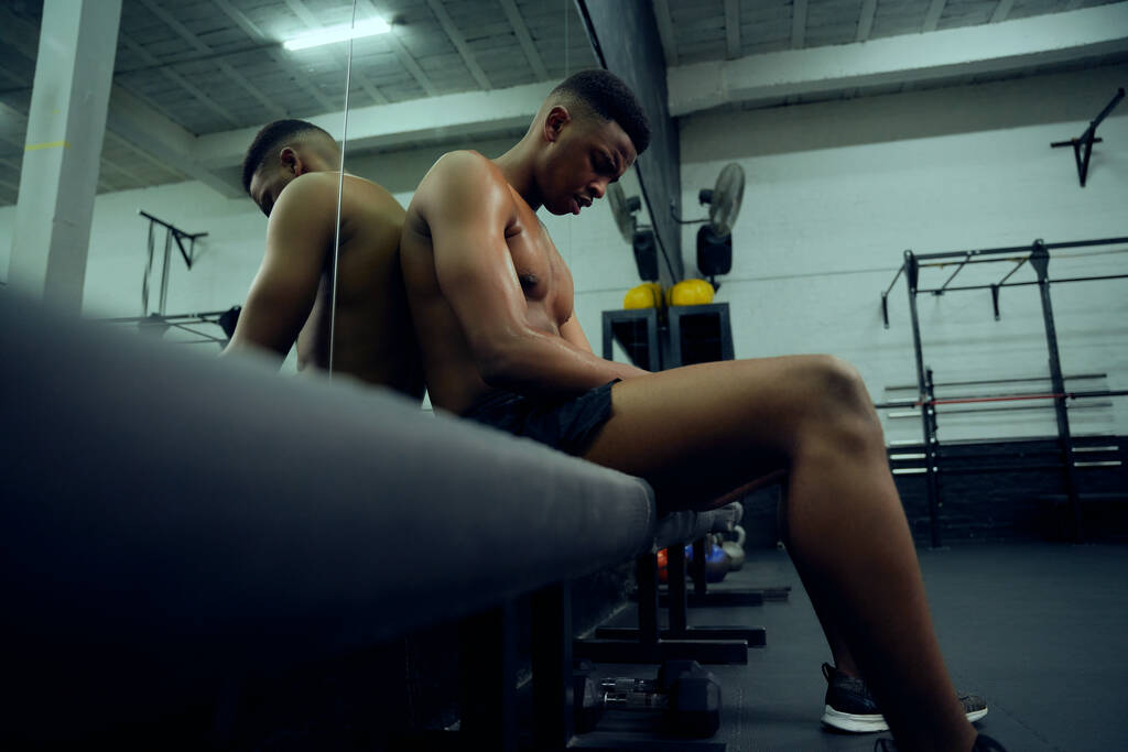 Jovem atleta afro-americano descansando no ginásio depois de um duro treinamento cruzado. Sem camisa mestiço mestiço macho sentado após o exercício. Foto de alta qualidade - Foto, Imagem