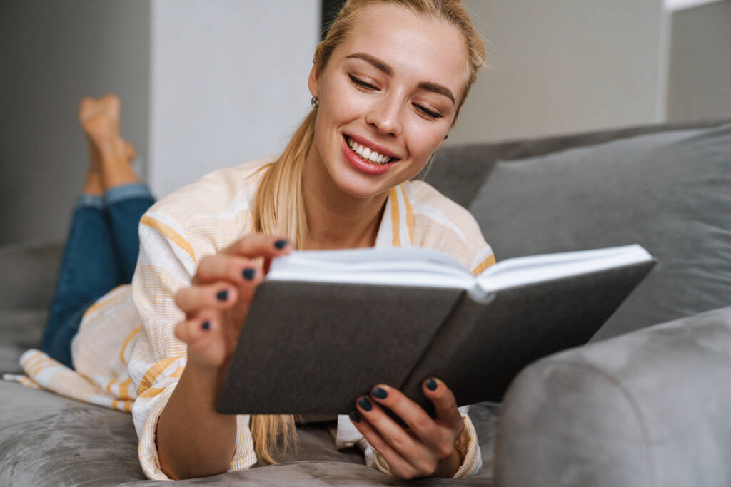 Onnellinen nainen hymyilee ja lukee kirjaa makatessaan sohvalla kotona - Valokuva, kuva